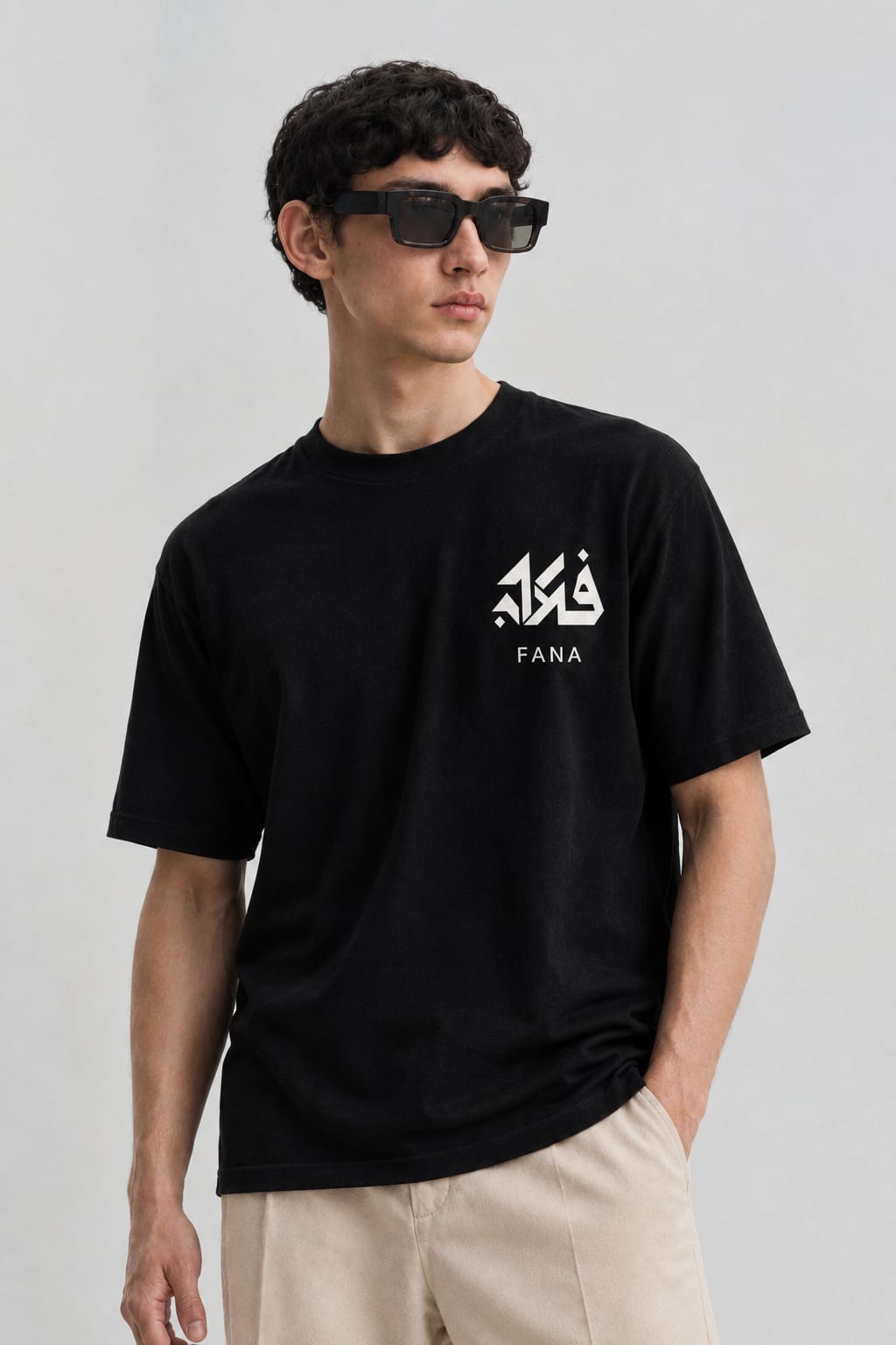 A black FANA tee with the فنا mark on the chest, worn with light trousers